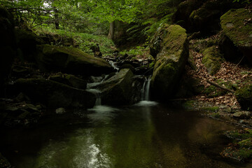 Ein idyllischer, schattiger Waldplatz mit einem sanft pl&auml;tschernden Wasserfall, der zwischen moosbewachsenen Felsen in einen ruhigen Teich flie&szlig;t &ndash; pure Naturidylle.