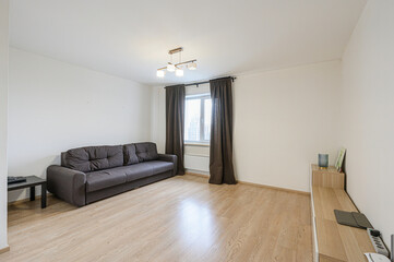 simple living room with a brown sofa, wooden floors, a window with dark curtains, and a light fixture