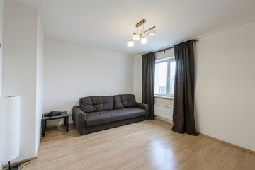 Bright room featuring a brown sofa, wooden floor, white walls, and window with brown curtains. Simple and modern interior design