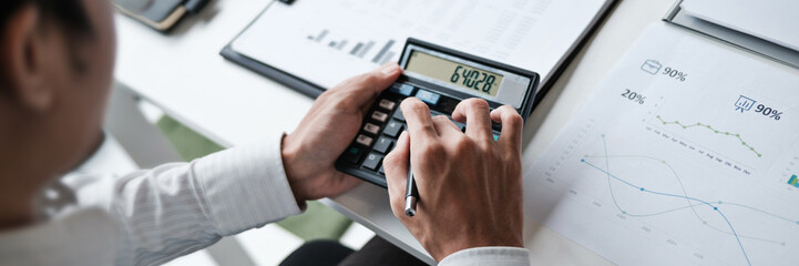 Focused businessman checking reports and verifying office documents with calculator and paper files...