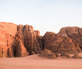 Fototapeta premium Scenic desert cliffs at sunset with red sandstone formations and sandy ground. Majestic rocky landscape with warm light, clear sky, and natural geological textures