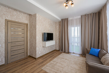 Cozy living room with beige walls, wooden floors, a TV, brown sofa, and soft rug. Natural light from the window with curtains