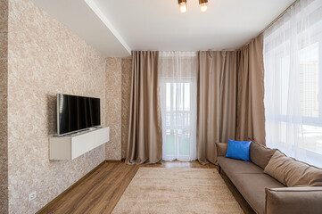 cozy living room features a wall-mounted TV, a light-colored patterned wall, neutral-toned curtains, a couch with a blue pillow, and a plush rug