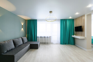 modern living room with a gray sofa, light wood floors, and teal curtains. A window provides natural light. The room has a minimalist aesthetic