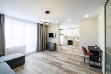 modern living room with open kitchen. Features include a gray sofa, wooden floor, TV, and stylish lighting. The kitchen has white cabinets and patterned backsplash