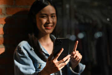 A Close-up of a young woman gestures while using her smartphone, standing by a brick wall with a bright smile