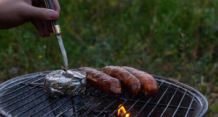 Grilled pork sausage and camambert on fire on a summer evening, at a grill party in a backyard. A hand is holding a pocket knife. Horizontal photo. Version 9.