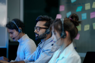 A diverse team of customer support agents works a late shift in a modern call center. Focused...