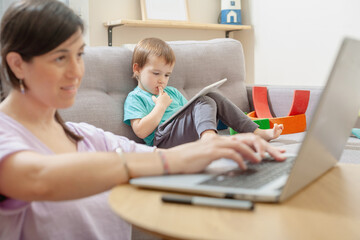 Mother working at home while toddler uses tablet on sofa
