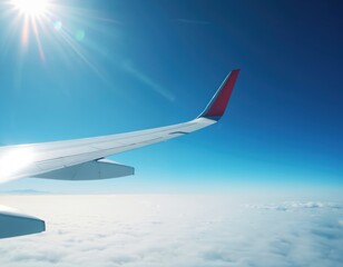 Fototapeta premium View from airplane window shows white wing and red tip against clear blue sky. Sun flares brightly in upper left corner. Fluffy white clouds form blanket below, with distant land visible on horizon.