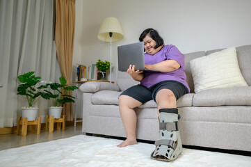 Asian woman wearing a leg brace sitting on sofa, holding laptop and smiling with excitement at home. Concept of disability, rehabilitation, healthcare, diversity, inclusion, and positive lifestyle.