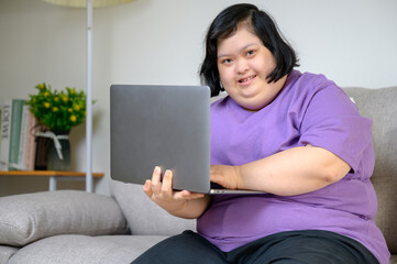 Asian woman with Down syndrome sitting on sofa and smiling while using a laptop indoors, wearing a...