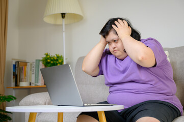 Asian woman with Down syndrome holding head with worried face while using laptop at home. Concept...