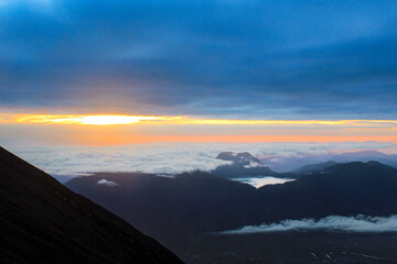 A breathtaking view of sunrise from a mountain summit, showing clouds blanketing the valley below and golden light spreading across the sky