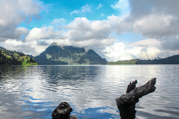 A tranquil tropical lake surrounded by forested mountains and dramatic clouds, with natural driftwood in the foreground and still water reflection