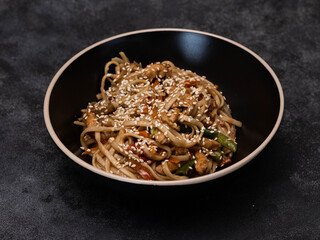 A black bowl with stirfried noodle, chicken meat and vegetables beautifully garnished with sesame seeds in a stylish bowl on black background.