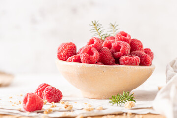 A rustic ceramic bowl filled with plump, fresh raspberries sits on a white table surrounded by scattered granola
