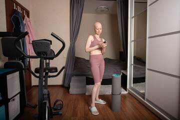 Bald woman with cancer in sportswear using smartphone before home fitness workout