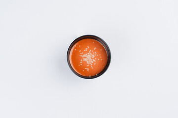 Overhead shot of a small bowl of orange sauce or dressing garnished with white sesame seeds, on a white background.