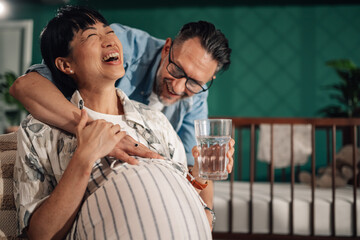 Happy pregnant woman laughing with husband holding glass of water in nursery