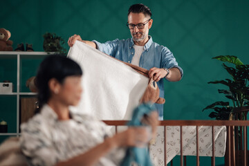 Father arranging baby blanket and teddy bear in crib, mother relaxing on sofa