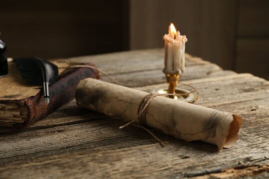Old paper scroll, book, feather and burning candle on wooden table, closeup
