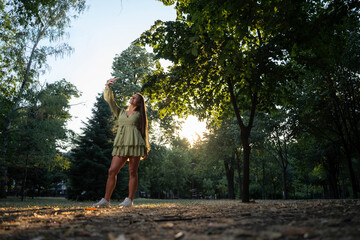 Fototapeta premium Happy beautiful young woman taking smiling selfie in park at golden hour sunset.
