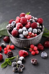 Mix of ripe frozen berries, leaves and bowl on dark textured table, closeup