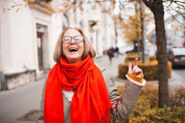 Smiling mature woman 55-60 year old wearing eye glasses, knitted red scarf and jacket in city...
