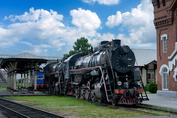An antique black steam train rests on the tracks. The locomotive is near an old brick building and a covered platform under a cloudy sky. Grass grows around the train's wheels