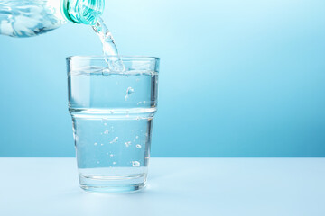 Pouring refreshing soda water from bottle into glass at white table against light blue gradient...