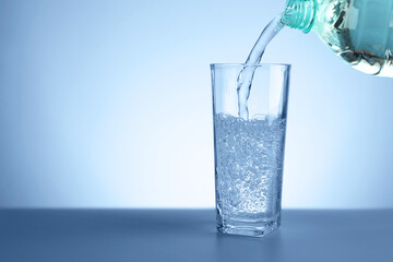 Pouring refreshing soda water from bottle into glass at table against light blue gradient background, closeup. Space for text