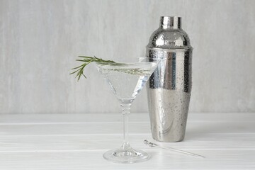 Martini cocktail with rosemary in glass and shaker on white wooden table, closeup