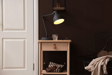 Wooden side table with glass of water, lamp and glasses near brown wall indoors