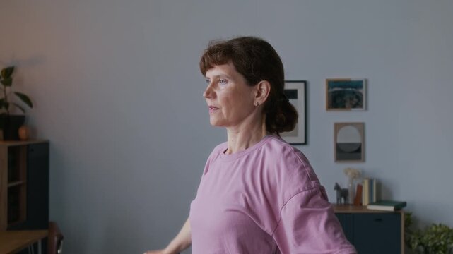Medium shot of elderly Caucasian woman in pink t-shirt doing repeats of lateral shoulder raise exercise using dumbbells at home, looking ahead with focused expression, concentrating on technique