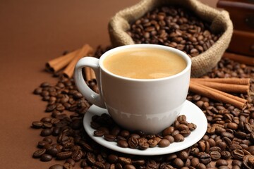 Cup of hot coffee, beans and cinnamon on brown background, closeup