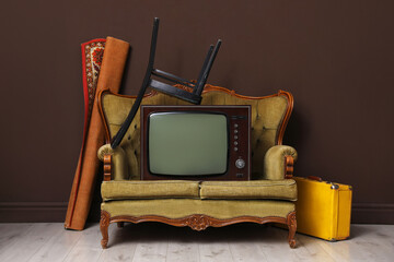 Retro TV on sofa, chair, suitcase and carpet near brown wall. Used stuff