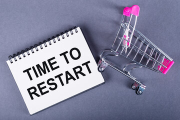 A notepad with the words TIME TO RESTART beside a miniature shopping cart on a gray background.