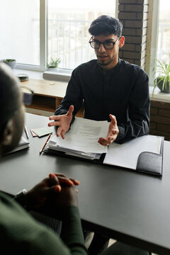 Vertical shot of serious young Hispanic man sitting at desk in front of his Black co-worker discussing new business plan