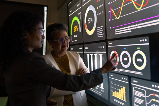 Asian female finance officer wearing glasses and senior shareholder standing and looking at real-time stock market analysis chart, investment consultant standing and presenting results in a conference - Powered by Adobe