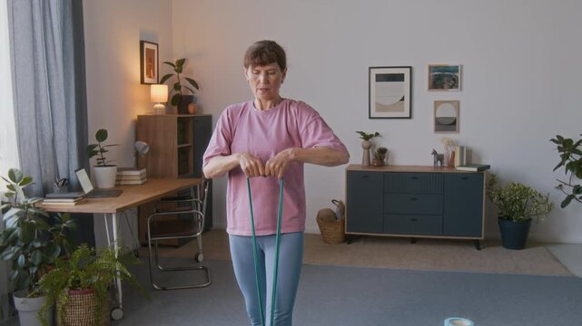 Medium tilt shot of fit elderly Caucasian woman in pink t-shirt, blue leggings performing back extension exercise, holding elastic resistance band, while doing training routine in living room at home