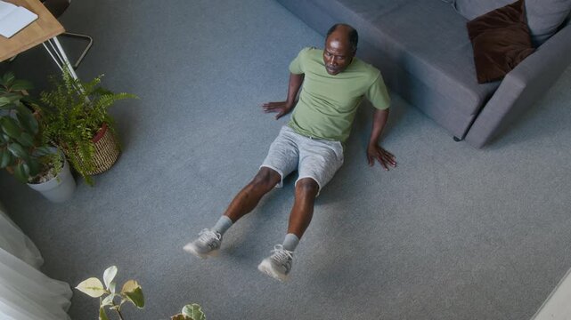 Full top view of athletic mature black man sitting on floor at home, performing repeats of abdominal scissor kicks exercise to strengthen body core muscles and develop endurance in old age