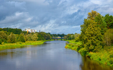Fototapeta premium A peaceful river with lush green banks, city buildings, and a bridge under dramatic cloudy skies.