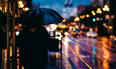 Person with umbrella waits for transport on rainy city street with bokeh lights © Mehebub