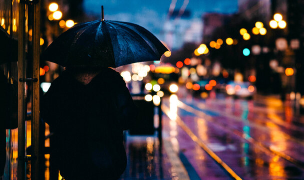 Person with umbrella waits for transport on rainy city street with bokeh lights