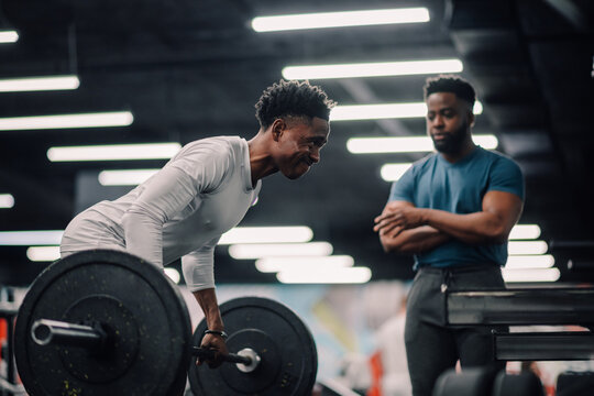 Determined male athlete lifting weights while personal trainer supervises in gym