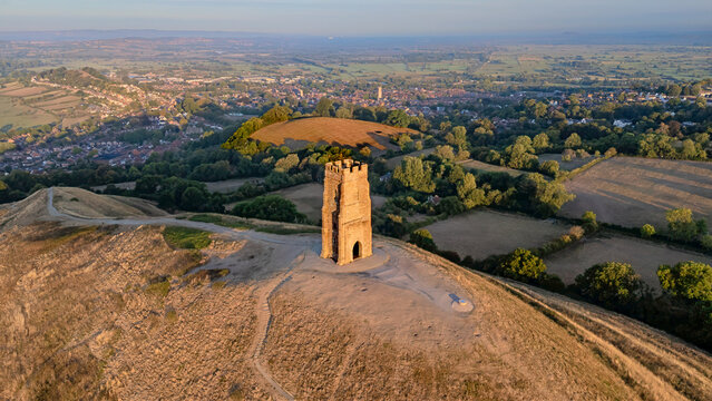 Drone photography of Glastonbury Tor with St Michael&rsquo;s Tower, overlooking the village of Glastonbury and the patchwork fields of Somerset. Captured at sunrise with warm golden tones.