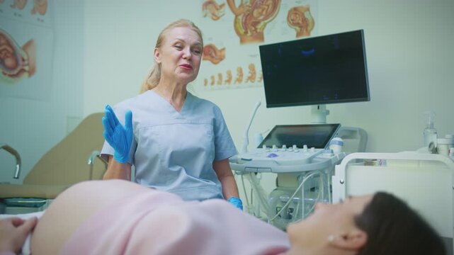 Senior female doctor in scrubs wearing gloves talking kindly to pregnant woman lying on bed. Specialist holding hands together while giving medical advice. Ultrasound equipment in clinic background.