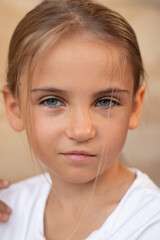 Young girl with long hair and blue eyes, wearing a white shirt, gazes confidently at the camera, showcasing her expressive features and natural beauty in a soft, warm light