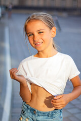 Young girl with blonde hair wearing a white shirt and denim shorts is playfully lifting her shirt,...